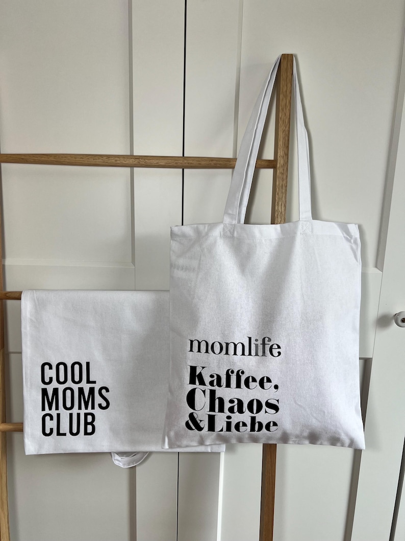 May include: White tote bag with black text that reads "momlife Kaffee, Chaos & Liebe" and a white tote bag with black text that reads "Cool Moms Club".