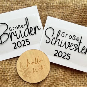 Könnte beinhalten: Zwei weiße Karten mit schwarzem Text. Der Text auf der oberen Karte lautet "Großer Bruder 2025". Der Text auf der unteren Karte lautet "Großer Schwester 2025". Ein kleiner Holz-Kreis mit dem Text "hallo Welt" befindet sich im Vordergrund.