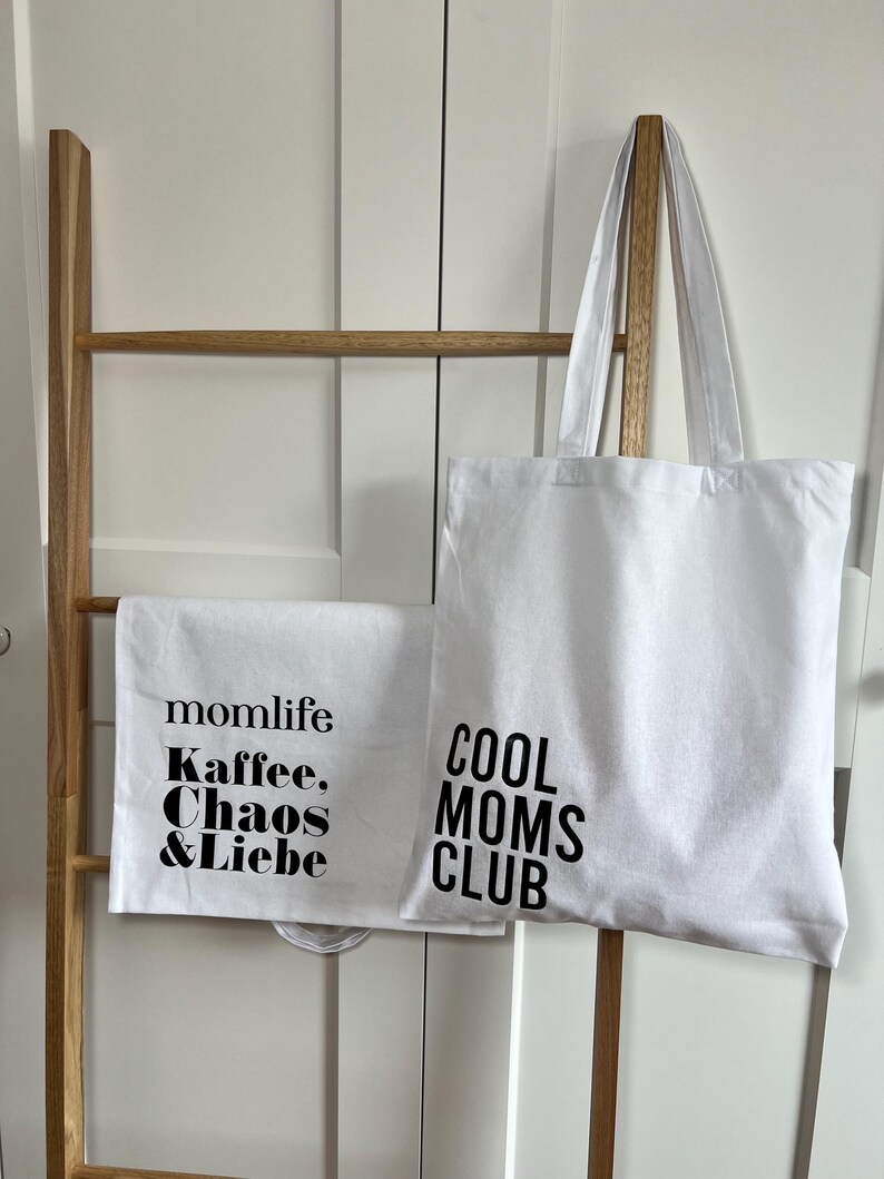 May include: Two white tote bags with black text. One bag says "momlife Kaffee, Chaos & Liebe" and the other says "COOL MOMS CLUB".