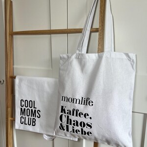 May include: Two white tote bags with black text. One bag says "Cool Moms Club" and the other says "momlife Kaffee, Chaos & Liebe".