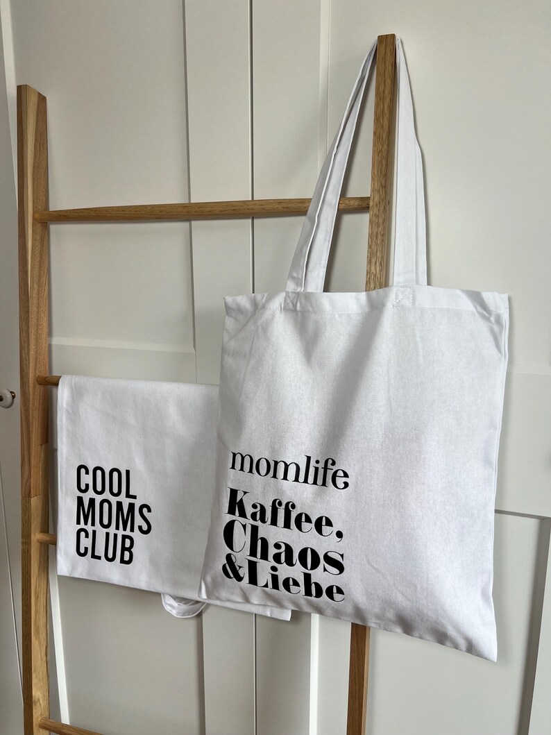 May include: Two white tote bags with black text. One bag says "Cool Moms Club" and the other says "momlife Kaffee, Chaos & Liebe".