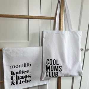 May include: Two white tote bags with black text. One bag says "momlife Kaffee. Chaos & Liebe" and the other says "Cool Moms Club".