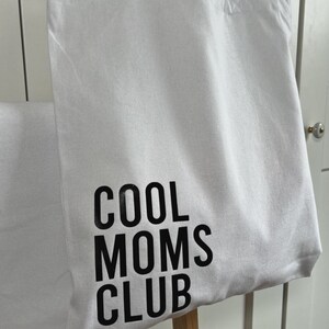 May include: White canvas tote bag with black text that reads "COOL MOMS CLUB".