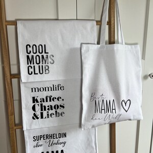 May include: Three white tote bags with black text. The top bag says "Cool Moms Club". The middle bag says "Momlife Kaffee, Chaos & Liebe". The bottom bag says "Superheldin ohne Umhang Mama". The tote bag in the middle has a heart on it and says "Beste Mama der Welt".