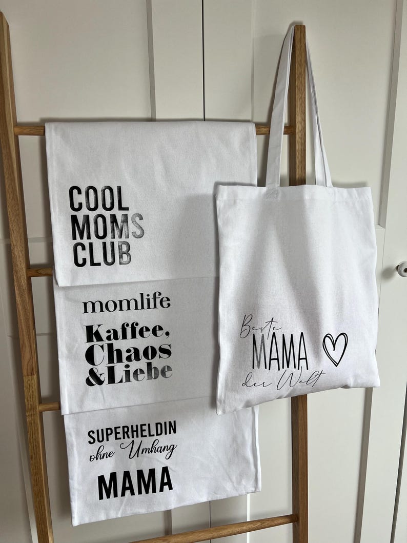 May include: Three white tote bags with black text. The top bag says "Cool Moms Club". The middle bag says "Momlife Kaffee, Chaos & Liebe". The bottom bag says "Superheldin ohne Umhang Mama". The tote bag in the middle has a heart on it and says "Beste Mama der Welt".