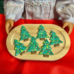 May include: A wooden tray holds six green Christmas tree-shaped cookies decorated with colorful dots. The cookies have brown bases and are arranged on a light-colored wooden tray, set against a red background.