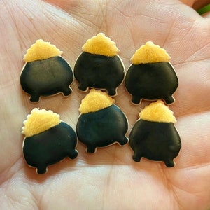 May include: Six black cauldron cookie decorations with gold glitter on top. The cauldrons have three legs and are decorated with gold glitter.