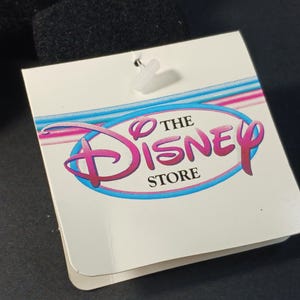 Op de afbeelding: Een wit label met de tekst "THE DISNEY STORE" in een roze en blauw logo. Het label is bevestigd aan een zwart object met een doorzichtige plastic sluiting. Het Disney-logo is een gestileerd schrift.