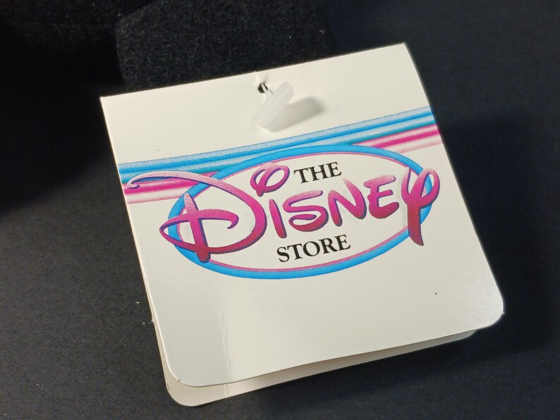 Op de afbeelding: Een wit label met de tekst "THE DISNEY STORE" in een roze en blauw logo. Het label is bevestigd aan een zwart object met een doorzichtige plastic sluiting. Het Disney-logo is een gestileerd schrift.