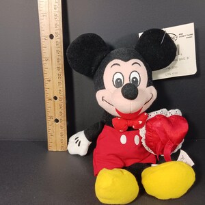 Op de afbeelding: Een Mickey Mouse pluche speelgoed met zwarte oren, een zwarte kop en een lachend gezicht. Mickey draagt een rode strik, een rode korte broek en gele schoenen. Hij houdt een rood hart met een witte kanten rand vast. Een houten liniaal ligt naast het speelgoed.