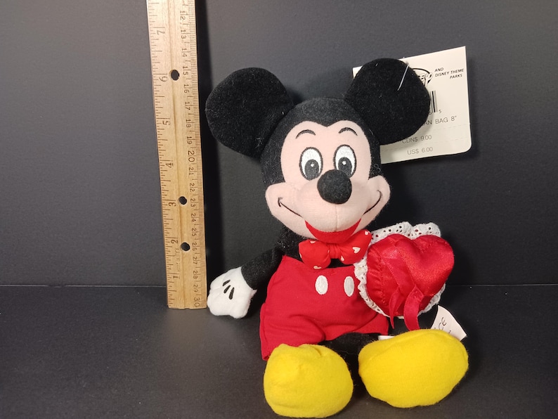 Op de afbeelding: Een Mickey Mouse pluche speelgoed met zwarte oren, een zwarte kop en een lachend gezicht. Mickey draagt een rode strik, een rode korte broek en gele schoenen. Hij houdt een rood hart met een witte kanten rand vast. Een houten liniaal ligt naast het speelgoed.
