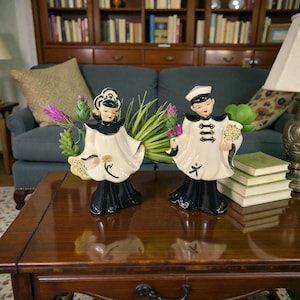May include: Two ceramic figurines, one female and one male, stand side-by-side on a wooden table. The figurines are dressed in white and black attire, holding flowers. The background includes a blue sofa, bookshelves, and a lamp.