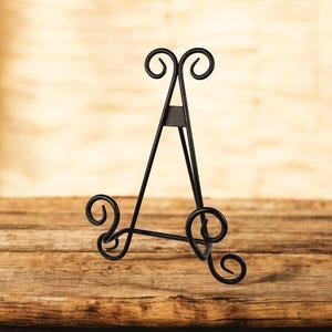May include: A black metal display easel with decorative scrollwork at the base and top. The easel is designed to hold a small picture frame or decorative item. The background is a blurred wooden surface.