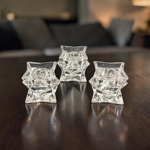 May include: Three clear glass star-shaped candle holders. Each holder has a faceted design and is sitting on a dark brown surface. The candle holders are arranged in a row, with the background out of focus.