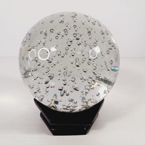 May include: A clear glass sphere filled with bubbles, resting on a black, geometric stand. The sphere is transparent, allowing light to pass through and highlighting the bubbles within. This decorative piece could be used as a paperweight or a unique home decor item.