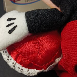 Op de afbeelding: Close-up van de hand en de korte broek van een Mickey Mouse pluche speelgoed. De witte handschoen heeft drie zwarte lijnen en de rode korte broek is afgezet met wit kant. De zwarte mouw is zichtbaar.