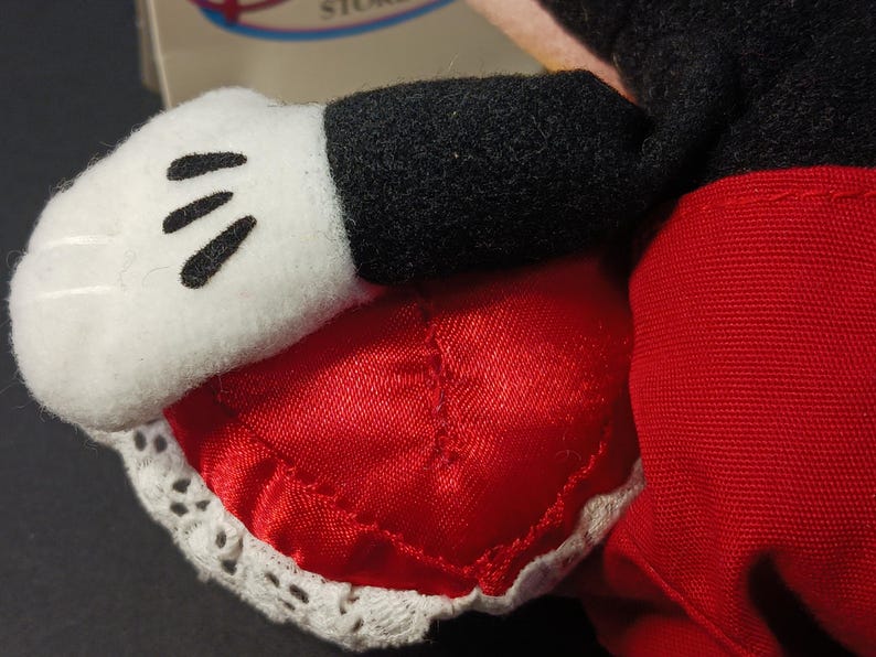 Op de afbeelding: Close-up van de hand en de korte broek van een Mickey Mouse pluche speelgoed. De witte handschoen heeft drie zwarte lijnen en de rode korte broek is afgezet met wit kant. De zwarte mouw is zichtbaar.