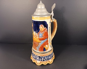 May include: A ceramic beer stein with a detailed relief scene of a man in a brown robe. The stein has a metal lid and handle, with a blue, white, and yellow color scheme. The base has a floral design.
