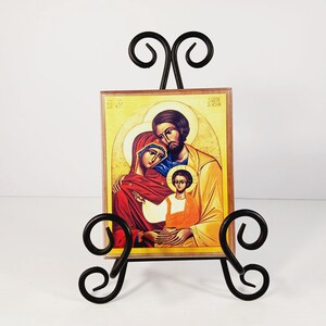 Op de afbeelding: Een icoon met een gezin, gepresenteerd op een zwarte smeedijzeren ezel. Het icoon toont een man, vrouw en kind met halo's, in rood, geel en bruin. De ezel heeft decoratieve krullen.