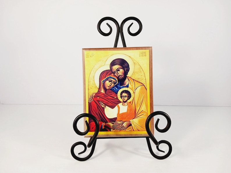 Op de afbeelding: Een icoon met een gezin, gepresenteerd op een zwarte smeedijzeren ezel. Het icoon toont een man, vrouw en kind met halo's, in rood, geel en bruin. De ezel heeft decoratieve krullen.