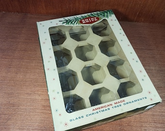 May include: A rectangular cardboard box for Shiny Brite glass Christmas tree ornaments. The box features a cream-colored exterior with a clear plastic window, a honeycomb-style internal divider, and red and green starburst patterns.