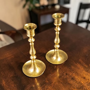 May include: Two vintage brass candlesticks. Each candlestick has a round base, a slender, ornate stem, and a cup-shaped top to hold a candle. The candlesticks are made of shiny, polished brass and are displayed on a dark wooden surface.