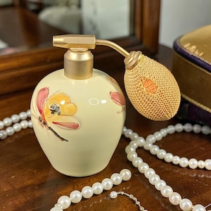 May include: A vintage perfume bottle with a cream-colored base and a gold-colored atomizer. The bottle is decorated with hand-painted butterfly designs in yellow, red, and black. A gold-colored beaded necklace is in the foreground.