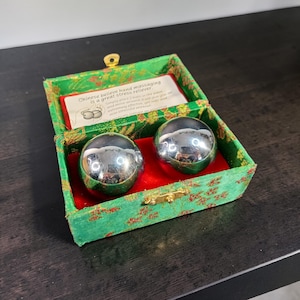 May include: A set of two shiny, chrome-colored massage balls inside a green and red decorative box. The box has a gold clasp and a printed label that reads "Chinese believe hand massaging is a great stress reliever."