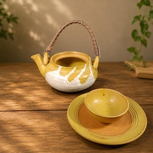 May include: A ceramic teapot with a mustard yellow and white glaze, featuring a woven brown handle. Beside it sits a matching mustard yellow bowl resting on a circular saucer with a terracotta-colored center and a yellow rim.