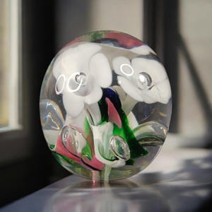 May include: A clear glass paperweight with white, pink, green, and blue floral inclusions. The paperweight is spherical and contains air bubbles, creating a unique decorative piece.