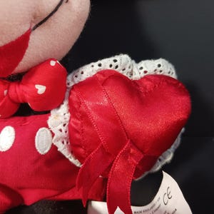 Op de afbeelding: Een Mickey Mouse-knuffel met een rode strik met witte hartjes en een rood hartvormig kussen met witte kanten rand. De pop draagt een rode outfit met witte knopen.