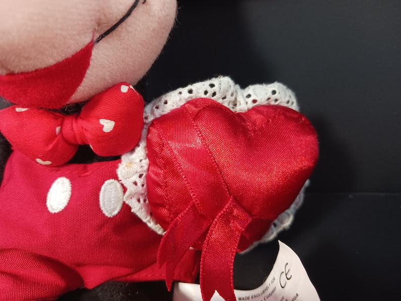 Op de afbeelding: Een Mickey Mouse-knuffel met een rode strik met witte hartjes en een rood hartvormig kussen met witte kanten rand. De pop draagt een rode outfit met witte knopen.
