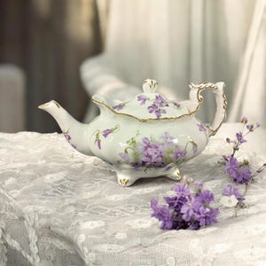 May include: A white porcelain teapot with a lid, adorned with purple floral patterns and gold trim. The teapot has a curved spout and handle, resting on three small feet. The design is delicate and elegant, suitable for serving tea.