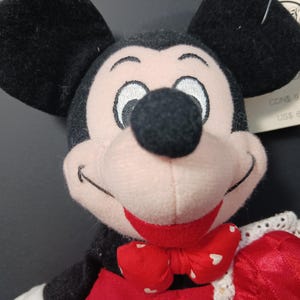 Op de afbeelding: Een Mickey Mouse knuffel met zwarte oren, een zwarte neus en een rode strik met witte hartjes. De pop heeft een roze gezicht en een rode outfit. Het label luidt "AN BAG 8".