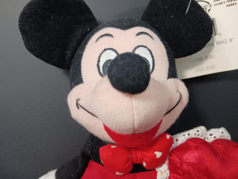 Op de afbeelding: Een Mickey Mouse knuffel met zwarte oren, een zwarte neus en een rode strik met witte hartjes. De pop heeft een roze gezicht en een rode outfit. Het label luidt "AN BAG 8".