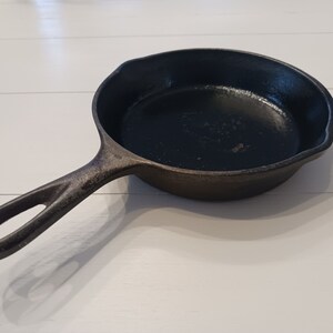 May include: A black cast iron skillet with a round cooking surface and a single handle featuring a teardrop-shaped hole at the end. The pan has a textured, dark finish and a small pour spout on the side, showing a sturdy, heavy-duty construction.