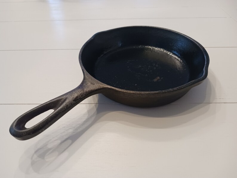 May include: A black cast iron skillet with a round cooking surface and a single handle featuring a teardrop-shaped hole at the end. The pan has a textured, dark finish and a small pour spout on the side, showing a sturdy, heavy-duty construction.