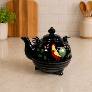 May include: A black ceramic teapot featuring a hand-painted rooster design in yellow, red, and green. The teapot has a rounded body, a curved spout, a matching lid with a small knob, and three short feet. It includes a metal handle attachment.