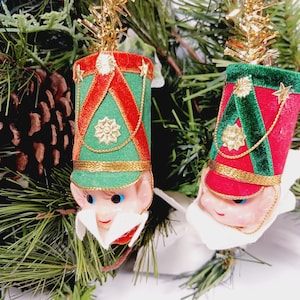 May include: Two Christmas tree ornaments shaped like toy soldiers. Each ornament has a red and green hat with gold trim and a smiling face. The ornaments are hanging on a Christmas tree with pine needles and a pine cone.
