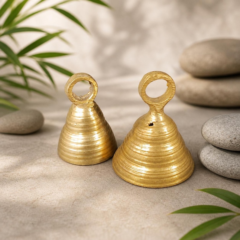 Puede incluir: Dos objetos dorados en forma de campana con un anillo en la parte superior. Las campanas tienen un dise&ntilde;o acanalado y se asientan sobre una superficie neutra con piedras lisas y vegetaci&oacute;n en el fondo. Las campanas son probablemente decorativas.