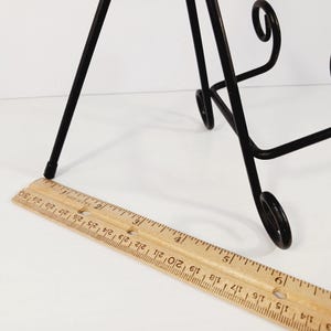 Op de afbeelding: Een zwartmetalen display-ezel met decoratief krulwerk. Een houten liniaal is naast de ezel geplaatst en toont metingen in centimeters. De ezel is ontworpen om een foto of bord te bevatten.