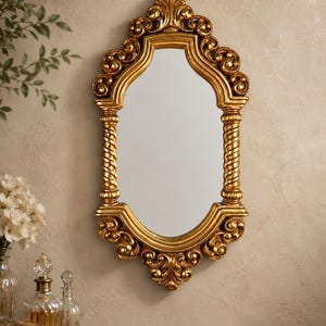 May include: A wall-mounted mirror featuring an ornate gold frame with intricate scrollwork at the top and bottom, and two vertical side columns with a twisted rope design. The mirror has a central glass pane with a curved, multi-faceted shape.