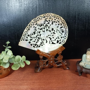 May include: A white mother-of-pearl fan with intricate cut-out floral and bamboo patterns, displayed on a dark brown carved wooden stand. Beside it sits a small green and white figurine on a wooden base and a copper-colored metal pot with heart cut-outs.