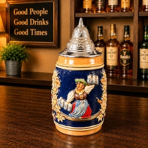 May include: A decorative beer stein with a silver lid and a detailed painted design. The stein features a blue background with a depiction of a woman holding beer steins. The stein is on a wooden surface.