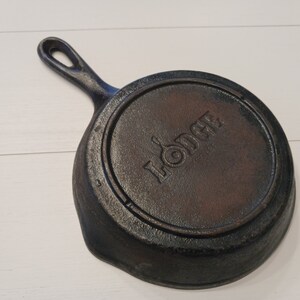 May include: A round black cast iron skillet with a single handle. The underside features the embossed Lodge logo in the center, which includes a small skillet icon within the letter O. The surface has a textured, dark metallic finish.