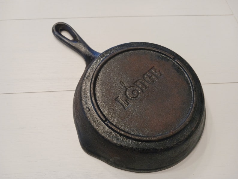 May include: A round black cast iron skillet with a single handle. The underside features the embossed Lodge logo in the center, which includes a small skillet icon within the letter O. The surface has a textured, dark metallic finish.