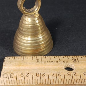 Puede incluir: Una peque&ntilde;a campana de lat&oacute;n antiguo con un asa de bucle. La campana tiene forma c&oacute;nica con crestas horizontales. Una regla de madera con medidas en pulgadas est&aacute; al lado de la campana.