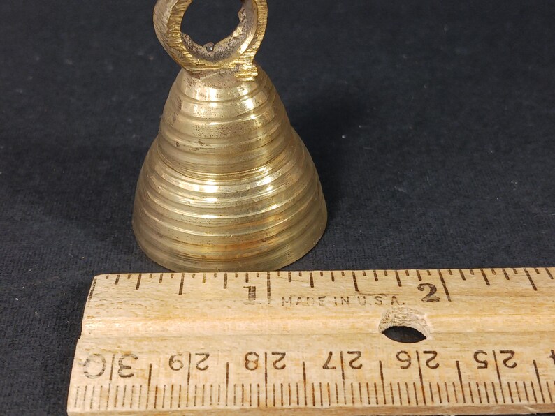 Puede incluir: Una peque&ntilde;a campana de lat&oacute;n antiguo con un asa de bucle. La campana tiene forma c&oacute;nica con crestas horizontales. Una regla de madera con medidas en pulgadas est&aacute; al lado de la campana.