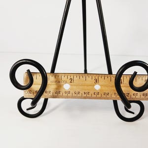 Op de afbeelding: Een zwart metalen tafel-ezel met een houten liniaalbasis. De ezel heeft een driepootontwerp en decoratief krulwerk. De liniaal is gemarkeerd in centimeters, tot 15 cm.