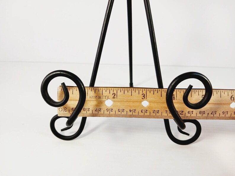Op de afbeelding: Een zwart metalen tafel-ezel met een houten liniaalbasis. De ezel heeft een driepootontwerp en decoratief krulwerk. De liniaal is gemarkeerd in centimeters, tot 15 cm.